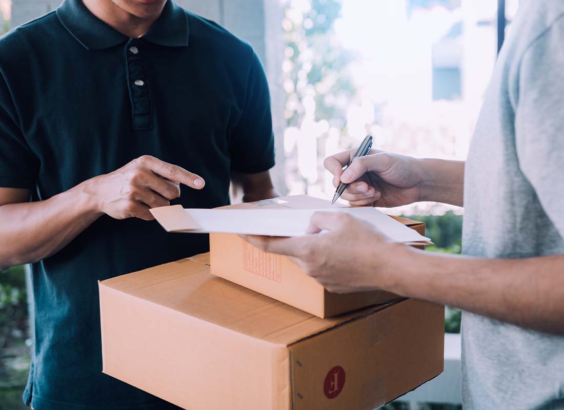 Parcel Delivery Contractor Insurance - Young Delivery Man Standing at the Door of a Home and Carrying Parcels for Young Male Signing for Delivery